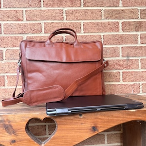 May include: A brown leather briefcase with a textured surface, a top handle, and an adjustable shoulder strap with a padded section. A thin, closed black laptop is positioned horizontally in front of the briefcase on a wooden surface.