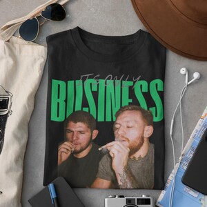 May include: Black t-shirt with the text "IT'S ONLY BUSINESS" in green block letters. The shirt features a photo of two men smoking. Other items in the image include a camera, sunglasses, and a tote bag.