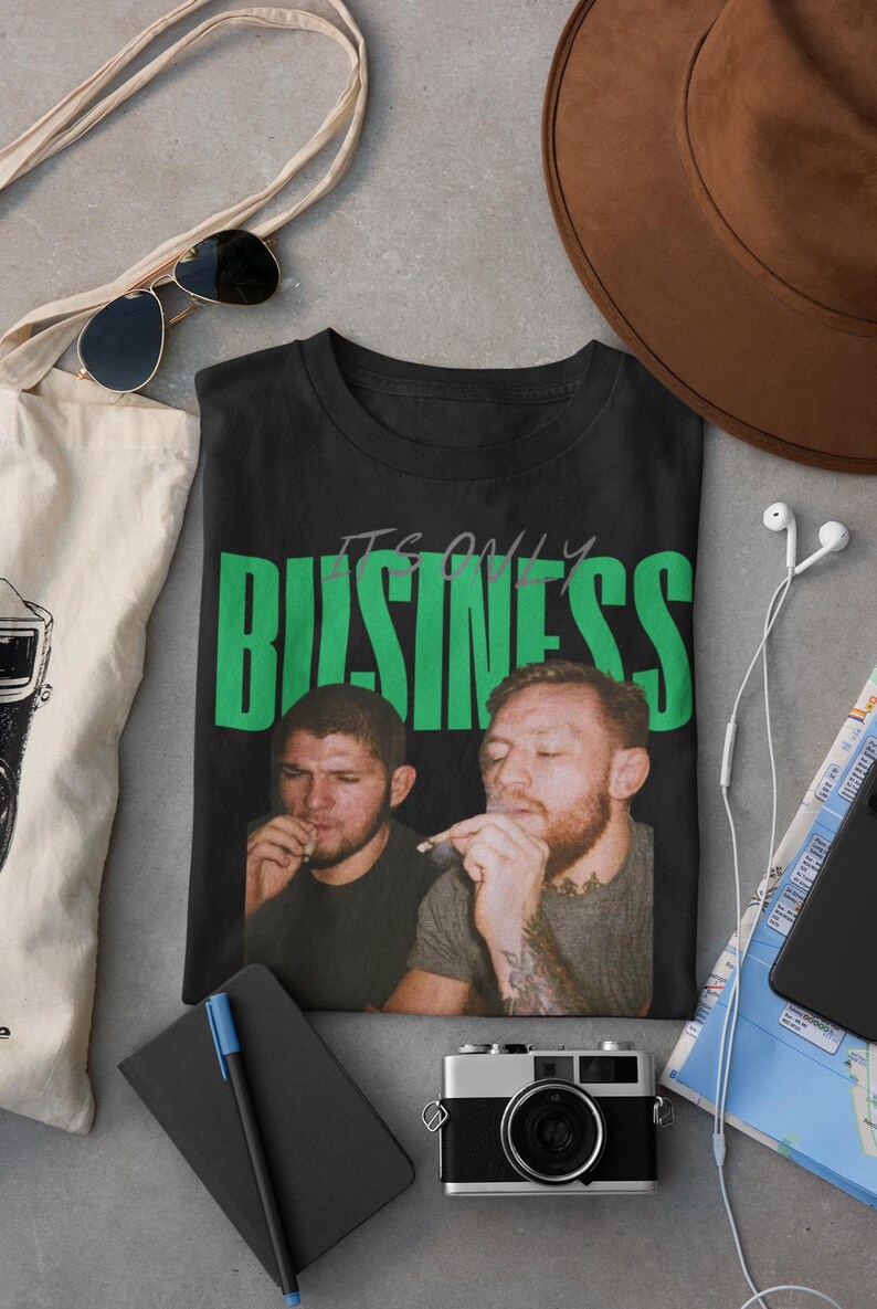 May include: Black t-shirt with the text "IT'S ONLY BUSINESS" in green block letters. The shirt features a photo of two men smoking. Other items in the image include a camera, sunglasses, and a tote bag.