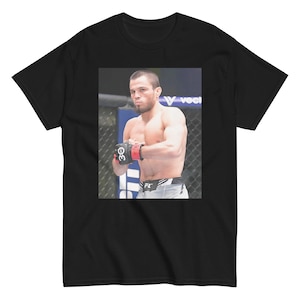 Puede incluir: Una camiseta negra con una foto de un hombre que lleva guantes de boxeo y un uniforme gris y negro. El hombre está en un ring de boxeo con un fondo azul y morado. El texto "UFC" es visible en el uniforme.