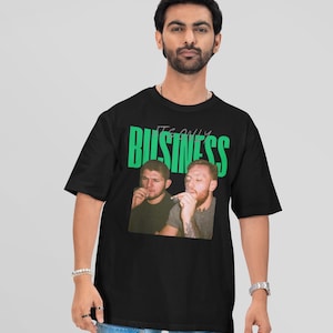 May include: Black t-shirt featuring a graphic of two men with cigarettes, with the text "IT'S ONLY BUSINESS" in green. The shirt is worn by a person with a beard, bracelets, and a watch.