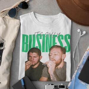 May include: White t-shirt with the text "IT'S ONLY BUSINESS" in green block letters. The shirt features a graphic of two men smoking. The shirt is surrounded by accessories including sunglasses, a hat, and a camera.