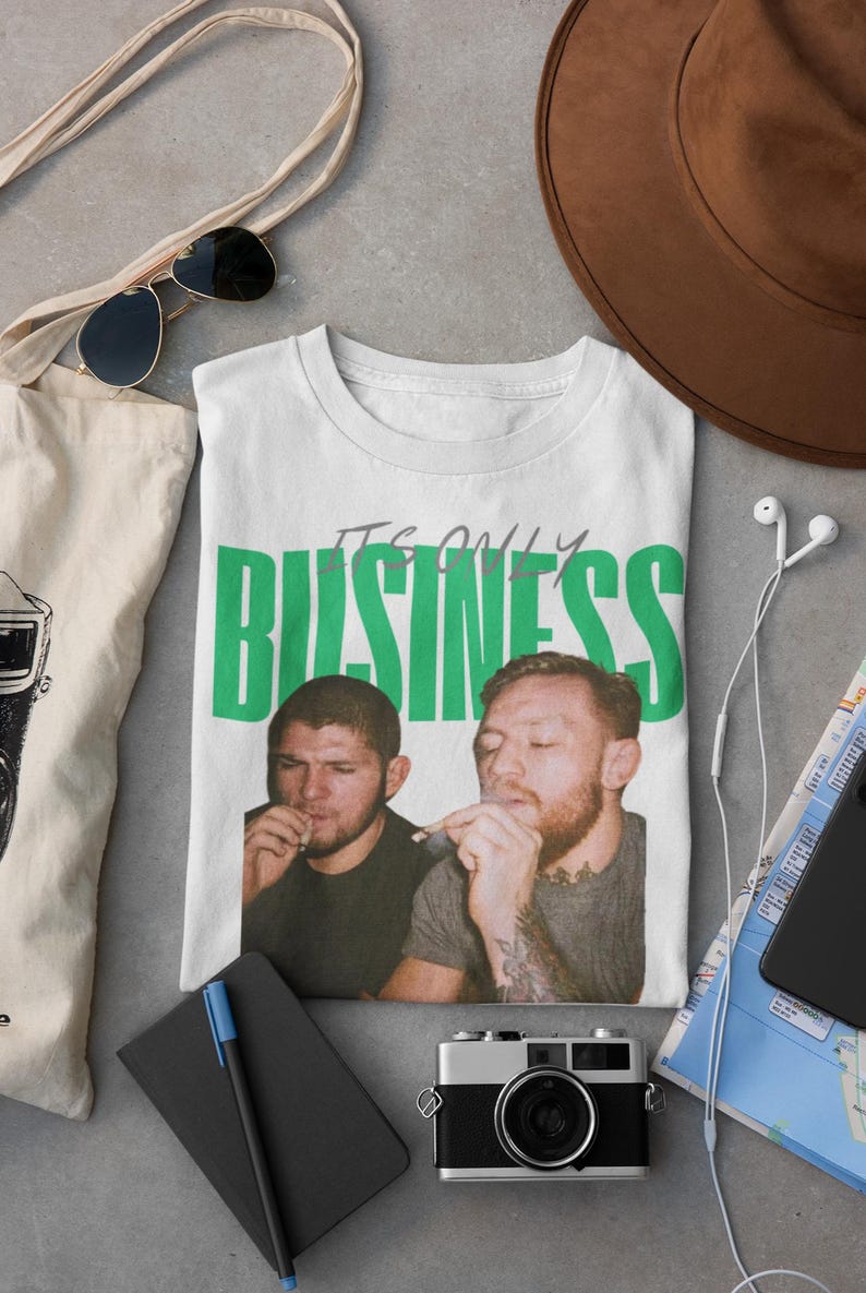 May include: White t-shirt with the text "IT'S ONLY BUSINESS" in green block letters. The shirt features a graphic of two men smoking. The shirt is surrounded by accessories including sunglasses, a hat, and a camera.