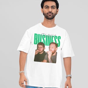 May include: White t-shirt with the green text "IT'S ONLY BUSINESS" and a photo of two men. The shirt is worn by a person with dark hair, wearing a watch and bracelets, and light blue jeans.