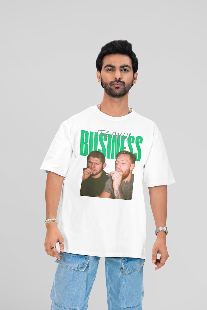 May include: White t-shirt with the green text "IT'S ONLY BUSINESS" and a photo of two men. The shirt is worn by a person with dark hair, wearing a watch and bracelets, and light blue jeans.