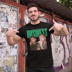 May include: Black t-shirt with the green text "IT'S ONLY BUSINESS" above a photo of two men. The man in the image is pointing at the shirt. The background includes posters and graffiti.