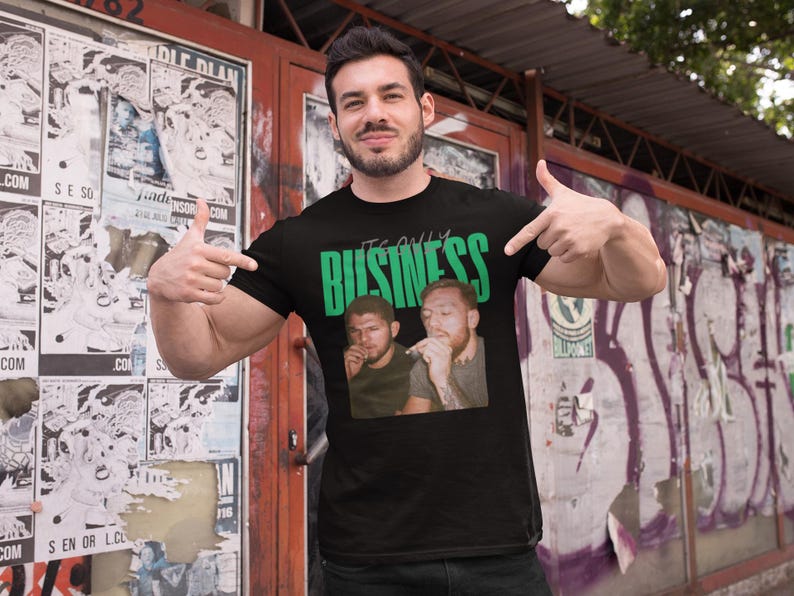 May include: Black t-shirt with the green text "IT'S ONLY BUSINESS" above a photo of two men. The man in the image is pointing at the shirt. The background includes posters and graffiti.