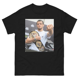 Puede incluir: Un hombre con una camisa blanca y pantalones cortos negros sostiene un cinturón de campeonato con una hebilla dorada y detalles en rojo, blanco y azul. El cinturón tiene las palabras "Bellator" y "Champion" en él.