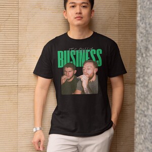 May include: Black t-shirt with a graphic of two men and the text "IT'S ONLY BUSINESS" in green. The t-shirt is worn by a person in light gray pants and a silver watch.