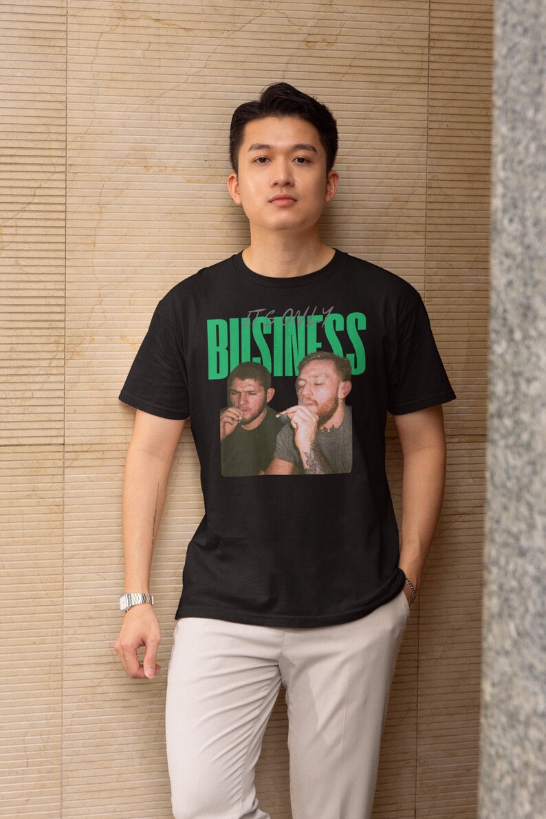 May include: Black t-shirt with a graphic of two men and the text "IT'S ONLY BUSINESS" in green. The t-shirt is worn by a person in light gray pants and a silver watch.