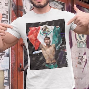 May include: A white t-shirt with a graphic of a man holding a Mexican flag. The man is wearing a black and white UFC fight shorts. The text "UFC" is visible on the shorts.
