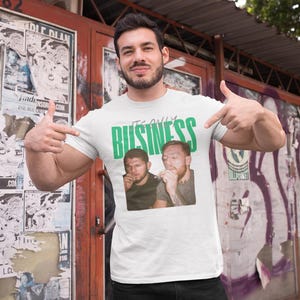 May include: White t-shirt with the text "IT'S ONLY BUSINESS" in green, featuring a graphic of two men. The person in the image is pointing at the shirt. The background includes posters and graffiti.
