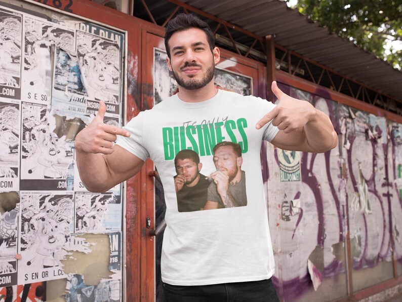 May include: White t-shirt with the text "IT'S ONLY BUSINESS" in green, featuring a graphic of two men. The person in the image is pointing at the shirt. The background includes posters and graffiti.