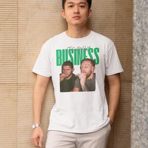 May include: White t-shirt with the text "IT'S ONLY BUSINESS" in green block letters. The shirt features a photo of two men. The person in the image is wearing a silver watch and light-colored pants.