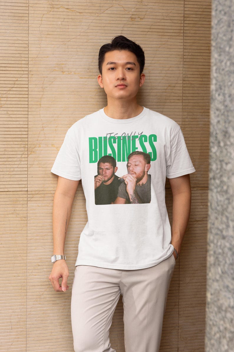 May include: White t-shirt with the text "IT'S ONLY BUSINESS" in green block letters. The shirt features a photo of two men. The person in the image is wearing a silver watch and light-colored pants.