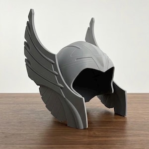 Könnte beinhalten: Ein mattgrauer Helm mit markanten, stilisierten Flügeln, die sich an beiden Seiten nach oben erstrecken. Der Helm hat eine glatte, abgerundete Kuppel mit geformten linearen Details und eine breite, offene Gesichtsöffnung aus festem Material.