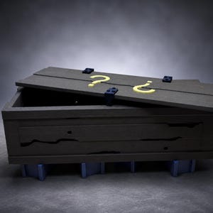 May include: A dark gray wooden crate with a partially open lid featuring two yellow question marks. The crate is secured with dark blue hinges and rests on four light blue block supports, showcasing a weathered texture with visible wood grain.