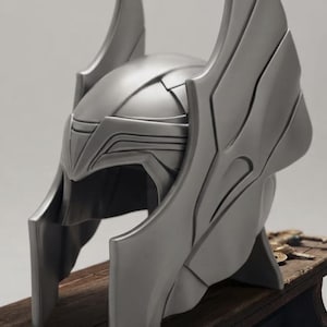 May include: A matte silver helmet featuring prominent, upward-curving wing-like structures on both sides. The helmet has a smooth, contoured surface with defined geometric lines across the brow and cheek guards, creating a structured, full-coverage design.