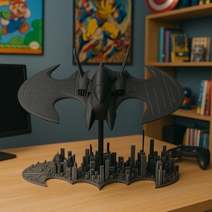 May include: A dark gray Batwing model, a replica of the iconic aircraft from the Batman universe. The model is perched above a cityscape base, with a detailed design. The background includes comic book art and a gaming controller.