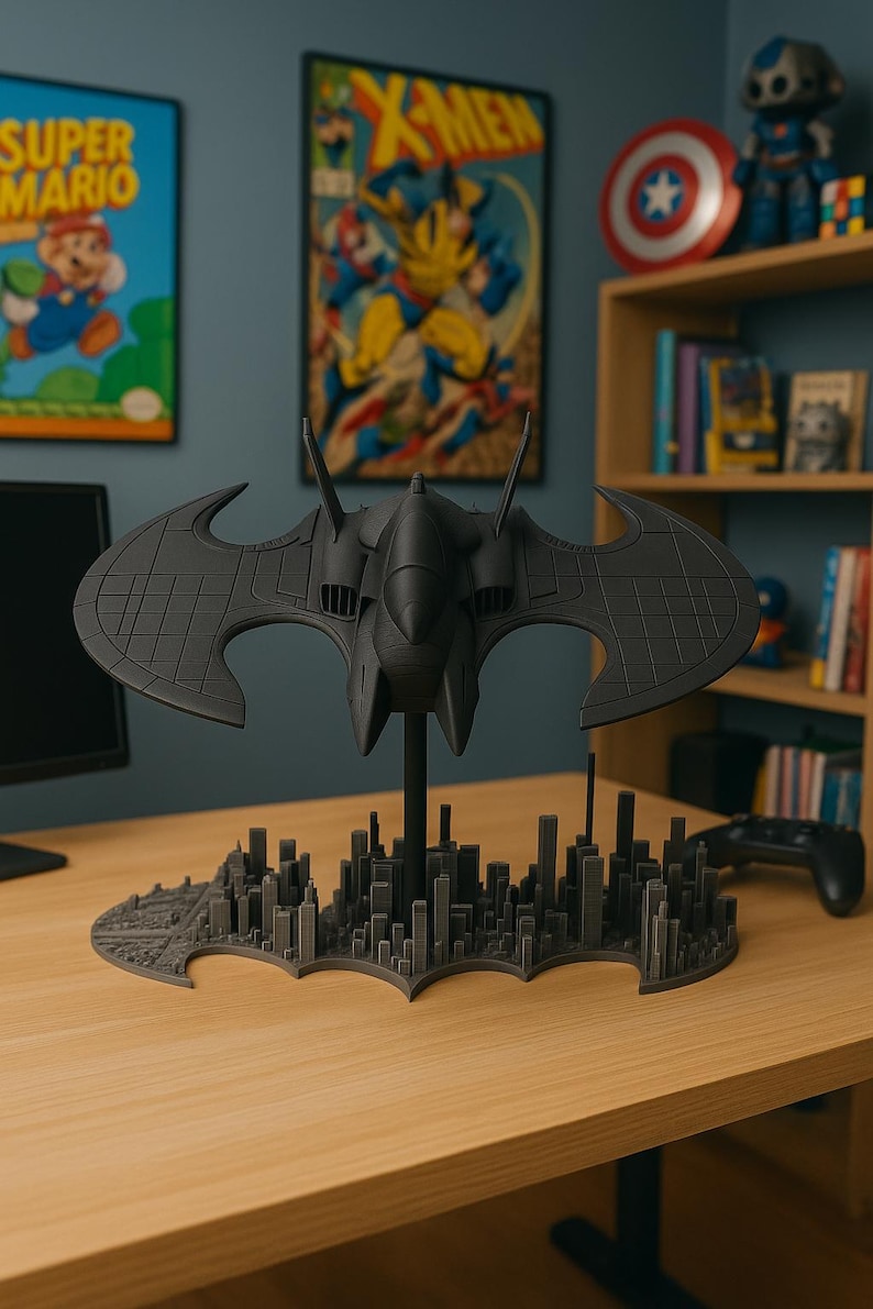 May include: A dark gray Batwing model, a replica of the iconic aircraft from the Batman universe. The model is perched above a cityscape base, with a detailed design. The background includes comic book art and a gaming controller.