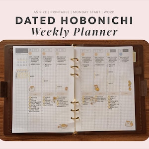 Puede incluir: Una agenda semanal Hobonichi fechada en tamaño A5 con inicio en lunes y diseño de semana en dos páginas. La agenda abierta presenta un diseño de cuadrícula con tareas escritas a mano, pegatinas amarillas y un mecanismo de anillas dorado.