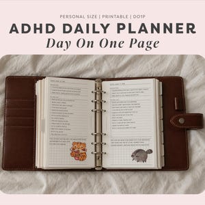May include: A brown leather ring-bound planner open to two pages labeled ADHD DAILY PLANNER Day On One Page. The pages feature sections for priorities and to-do lists, with a small tiger sticker and a raccoon sticker on the bottom.