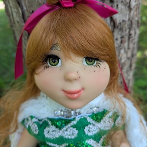 May include: A handcrafted doll with red hair and green eyes. The doll wears a green dress with white lace detailing and a white fur-trimmed cape. A pink bow adorns the doll's hair. The doll has freckles and a gentle expression.