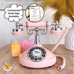Pu&ograve; includere: Un telefono rosa in stile vintage con quadrante rotante e dettagli argentati. Il telefono ha un cavo a spirale per il ricevitore e le parole "Leave a Message" sul quadrante. Un cartello "Audio Guest Book" &egrave; visibile sullo sfondo.