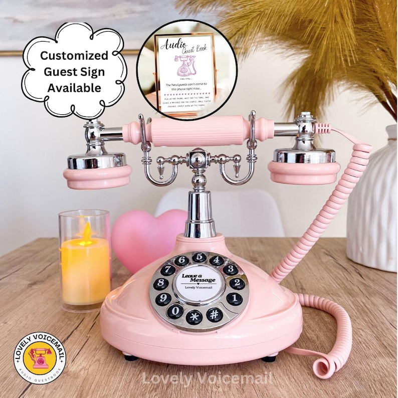 Pu&ograve; includere: Un telefono rosa in stile vintage con quadrante rotante e dettagli argentati. Il telefono ha un cavo a spirale per il ricevitore e le parole "Leave a Message" sul quadrante. Un cartello "Audio Guest Book" &egrave; visibile sullo sfondo.