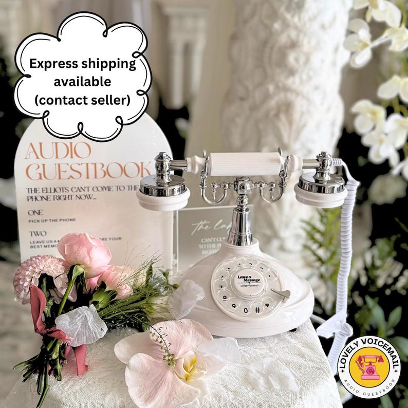 Pu&ograve; includere: Un telefono bianco in stile vintage con quadrante rotante e dettagli argentati. Il telefono fa parte di un'impostazione di libro degli ospiti audio, con un cartello che recita "Audio Guestbook". Sono visibili anche un bouquet di fiori rosa e un adesivo "Lovely Voicemail".