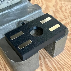 Puede incluir: Un componente de herramienta rectangular negro con una abertura circular central y cuatro placas met&aacute;licas rectangulares. El componente est&aacute; colocado sobre una base de metal, con una superficie de madera visible en el fondo.