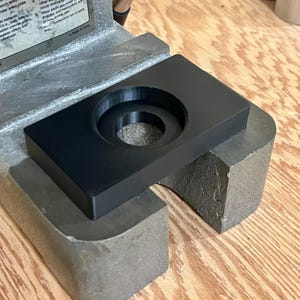 Puede incluir: Un componente de herramienta negro y rectangular con una hendidura circular. El componente est&aacute; colocado sobre una base de metal gris, probablemente parte de una m&aacute;quina o herramienta m&aacute;s grande. Vista de cerca.