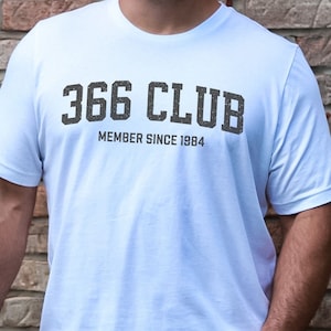 May include: White t-shirt with the text "366 CLUB MEMBER SINCE 1984" in a distressed, dark gray font. The shirt is short-sleeved and worn with blue jeans. The person is standing in front of a brick wall.