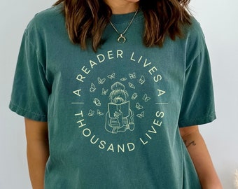 May include: Teal-colored t-shirt with the text "A Reader Lives A Thousand Lives" in a circular design. The design features a person reading a book surrounded by butterflies. The shirt is a comfortable, casual style.