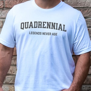 May include: White t-shirt featuring the text "QUADRENNIAL" and "LEGENDS NEVER AGE" in a dark, textured font. The short-sleeved shirt is worn by a person with their hand in their pocket. The background is a brick wall.