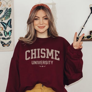 May include: A maroon sweatshirt with the text "CHISME UNIVERSITY" in white lettering. The sweatshirt has a relaxed fit and long sleeves.
