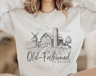 May include: A light gray sweatshirt with a black and white farm scene graphic, including a windmill, barn, and silo. The text "Old-Fashioned on purpose" is printed below the graphic. The sweatshirt is a crew neck style.