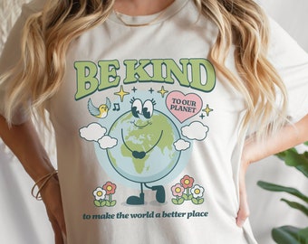 May include: White t-shirt with a green graphic that says "BE KIND" and features a cartoon Earth with a heart that says "TO OUR PLANET". The graphic also includes a bird, clouds, and flowers. The text below the graphic says "to make the world a better place."
