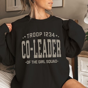 May include: Black sweatshirt with the text "TROOP 1234 CO-LEADER -OF THE GIRL SQUAD-" in a distressed, off-white font. The sweatshirt has a crew neck and long sleeves. The design includes star accents.