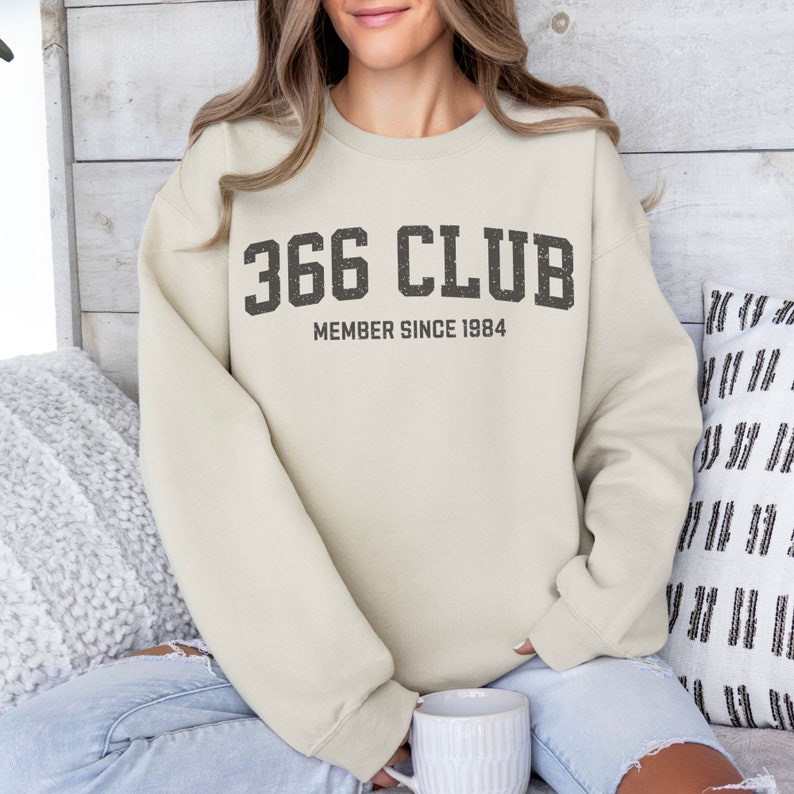 Op de afbeelding: Een beige sweatshirt met de tekst "366 CLUB MEMBER SINCE 1984" gedrukt op de voorkant.