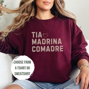 May include: A maroon sweatshirt with the text "TIA MADIRNA COMADRE" in white with a heart symbol between "TIA" and "MADIRNA". The text has a distressed look. The model is wearing the sweatshirt and blue denim shorts. The text "CHOOSE FROM A TSHIRT OR SWEATSHIRT" is written in white on a white circle with a black outline.