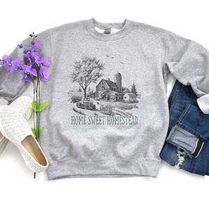 K&ouml;nnte beinhalten: Grauer Pullover mit einer Schwarzwei&szlig;illustration eines Bauernhauses, eines Autos und B&auml;umen. Der Text "HOME SWEET HOMESTEAD" ist unter der Illustration gedruckt.