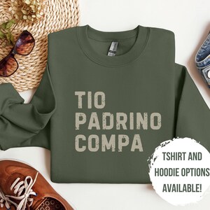 May include: A green sweatshirt with the text "TIO PADRINO COMPA" in a distressed white font. The text is centered on the sweatshirt. The sweatshirt is laid flat on a white surface. T-shirt and hoodie options are available.