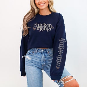 May include: A navy blue sweatshirt with the text "chicken mama" and a small chicken graphic on the front. The left sleeve has a list of names printed in brown: Ginger, Henrieta, Rosie, Goldie, Daisy, Blanch, Edith, Betty, Jill, Jane, Georgia, Dolly, Carrie, Reba, Rose.
