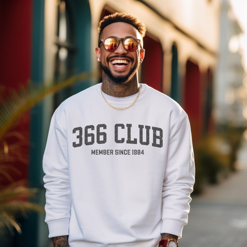 Op de afbeelding: Wit sweatshirt met zwarte tekst die "366 CLUB MEMBER SINCE 1984" luidt. Een man draagt het sweatshirt en glimlacht.
