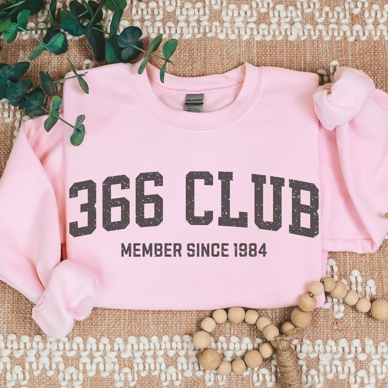 Op de afbeelding: Roze sweatshirt met de tekst "366 CLUB MEMBER SINCE 1984" in zwart.