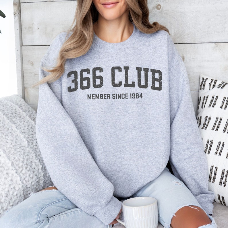 Op de afbeelding: Grijs sweatshirt met de tekst "366 CLUB MEMBER SINCE 1984" gedrukt op de voorkant.