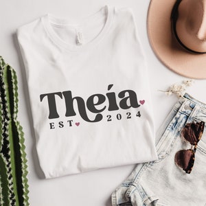 May include: White t-shirt with black text that reads "Theia EST 2024" with a small pink heart between "EST" and "2024".