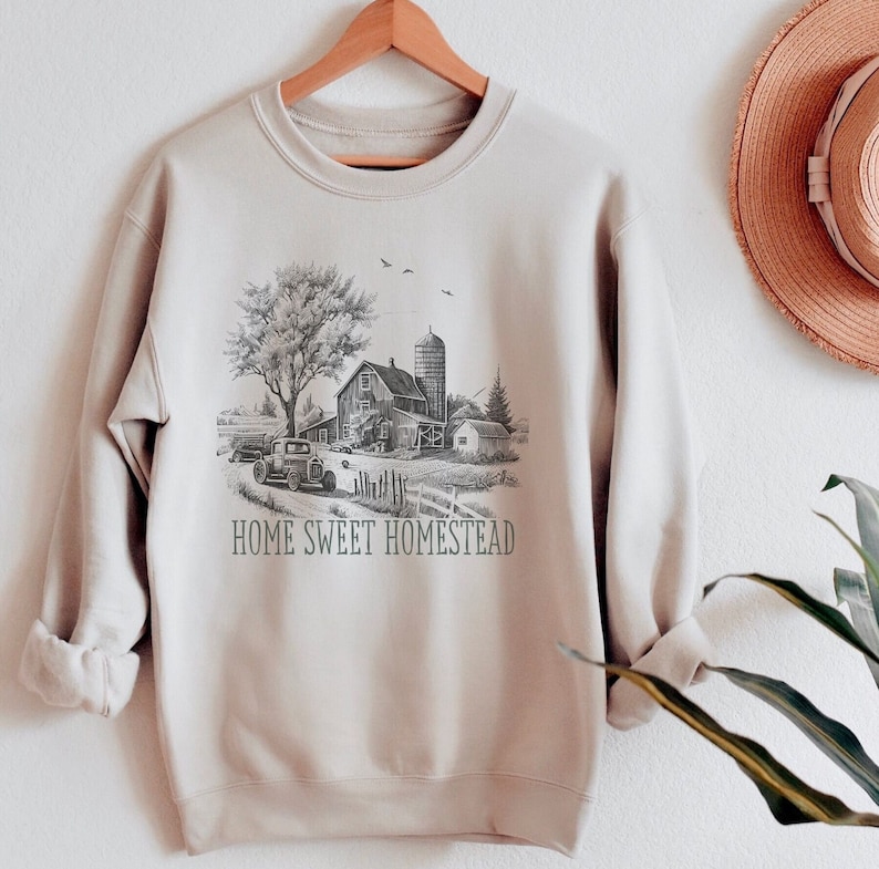 K&ouml;nnte beinhalten: Ein beiger Pullover mit einer Schwarzwei&szlig;illustration eines Bauernhauses, eines Autos und B&auml;umen. Der Text "Home Sweet Homestead" ist unter der Illustration gedruckt.