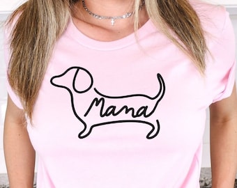 May include: A light pink short-sleeved t-shirt featuring a black line art graphic of a dachshund dog. The word "Mama" is written in a cursive script font within the body of the dog illustration. The shirt has a crew neckline and a knotted hem.